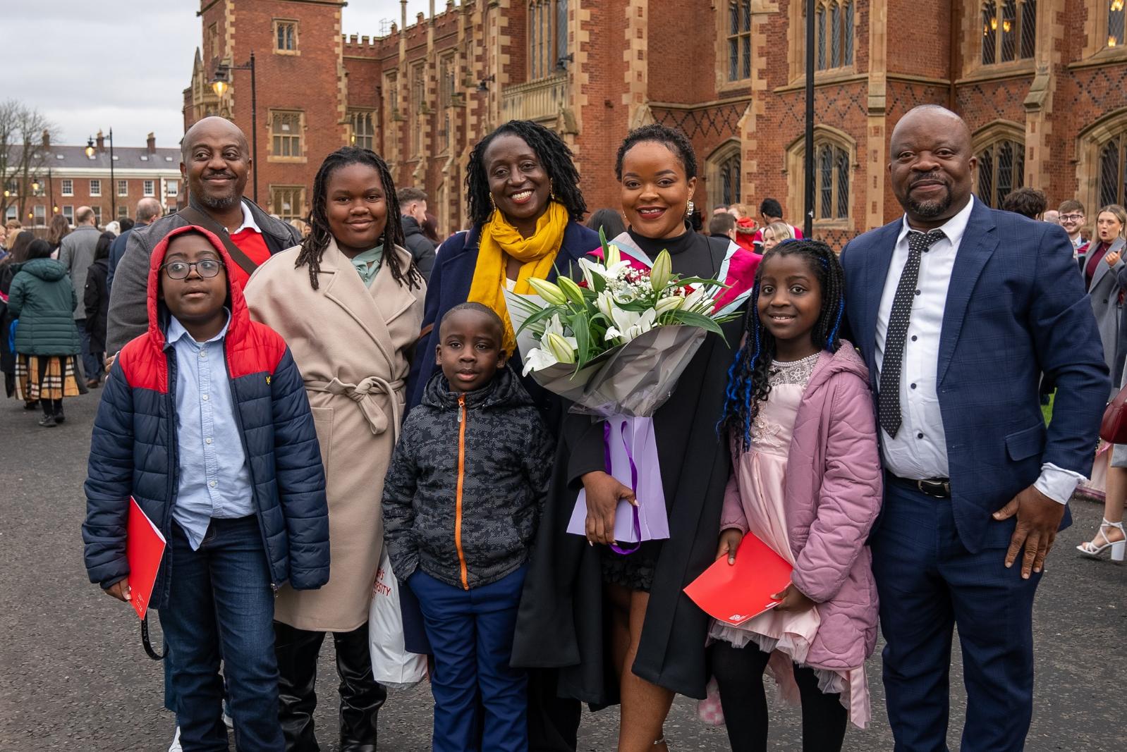 A graduate standing with their family outside a Queen's University Belfast building, celebrating their academic achievement.