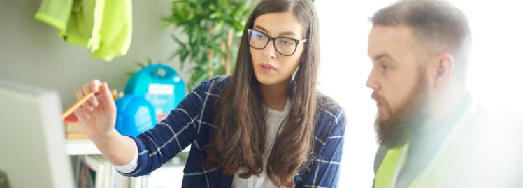 young woman wearing glasses showing something on a computer screen in an office to a man wearing a high-visibility jacket