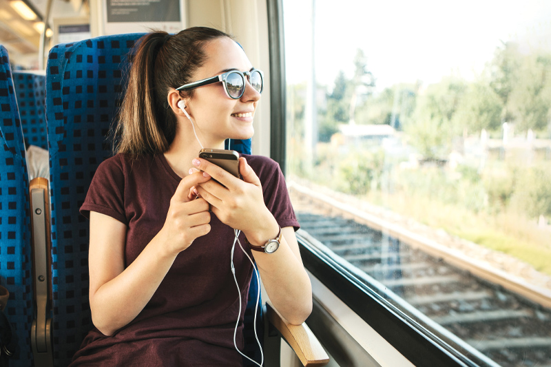 smiling young woman sitting on a train listening to something on her iPhone and looking out of the window